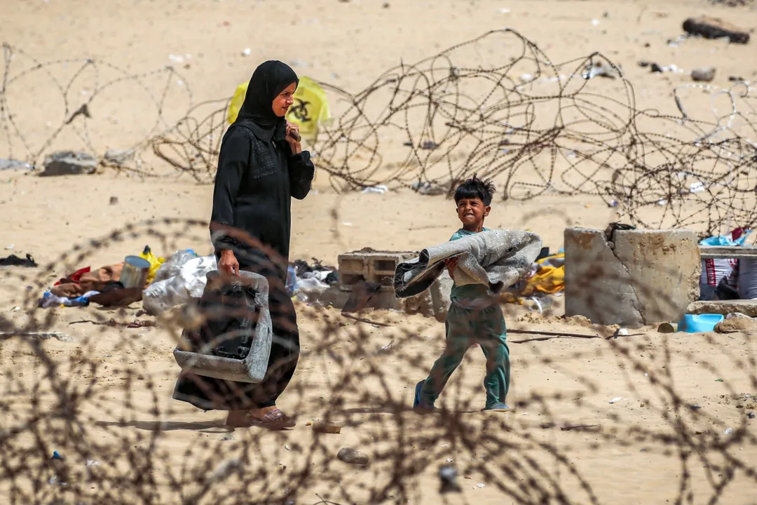 Uma mulher e um menino passam com pertences por cercas de arame farpado enquanto fogem de Rafah