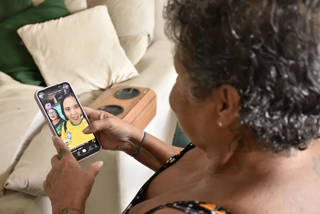 Emília Nunes na sala de sua casa usando o celular