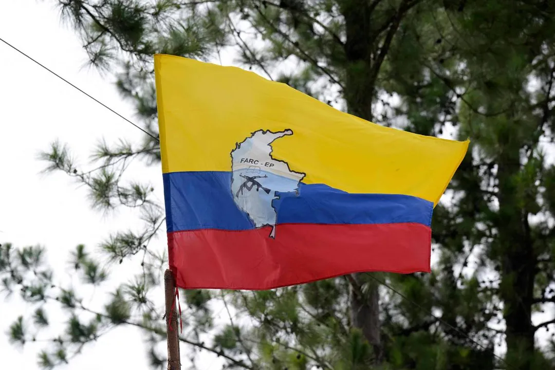 Bandeira com faixas horizontais amarela, azul e vermelha, hasteada em mastro diante de árvores. No centro, mapa da Colômbia em cinza com a sigla 'FARC-EP' e desenho de um fuzil preto.
Bandeira colombiana com o logotipo das Farc