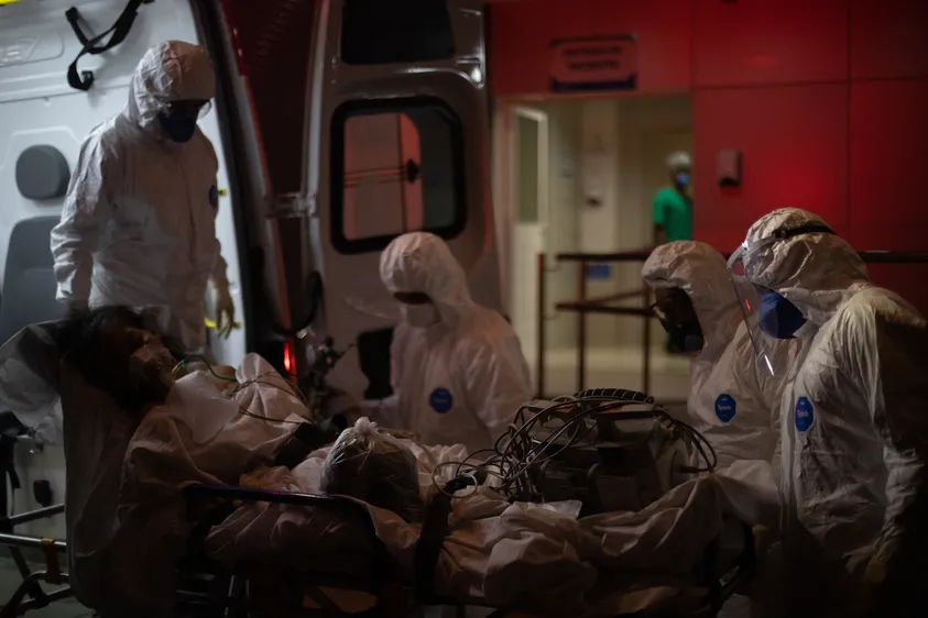 Médicos com equipamentos de proteção individual (EPIs), descendendo da ambulância para deixar paciente com covid-19 em hospital.