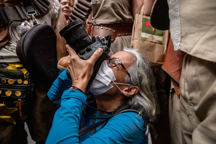 Na foto a fotojornalista Shirley Stolze em seu trabalho, durante o desfile de 2 de julho de 2023.
Essa foto representa o que há de melhor em minha profissão. Durante a apresentação dos cangaceiros de Ipitanga, o grupo fez uma roda e a fechou, com seus integrantes apontando suas armas e outros instrumentos figurativos para cima. Antecipando o gesto, Shirley pulou por baixo das pernas de um deles e, ajoelhada no chão, fez o que ela faz de melhor. Quem já acompanhou Shilóka, como carinhosamente a chamamos, em uma pauta, sabe o quanto ela é incansável e completamente apaixonada pelo que faz. Nós, é claro, somos igualmente apaixonados e inspirados por seu olhar.