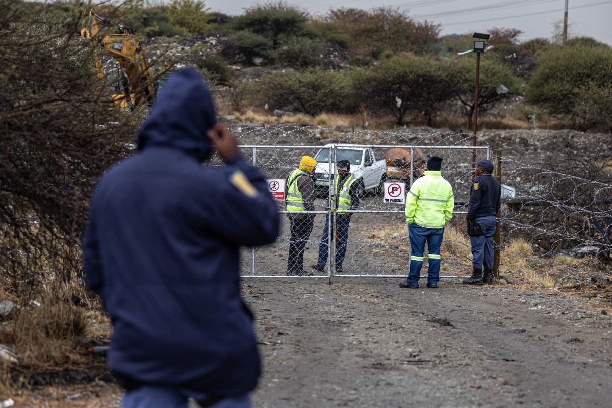 Integrantes da equipe de segurança conversam com policiais do Serviço de Polícia da África do Sul (SAPS) através de um portão, perto de um poço de mina em Nkaneng, nas imediações de Marikana, em 11 de agosto de 2026; no local, 14 garimpeiros ilegais foram encontrados mortos enquanto operações de resgate estão em andamento.