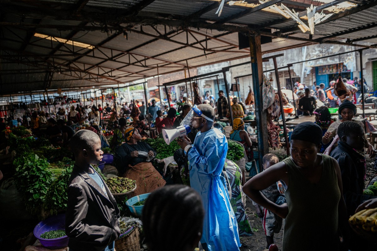 Irenge Biringanine Prince, líder da comunidade "U Report Goma", vestindo equipamento de proteção, conscientiza comerciantes ao lado de jovens membros do grupo "U Report Goma", que dão continuidade às atividades de conscientização no mercado Alanine utilizando megafones e discussões em grupos focais, como parte das medidas de prevenção e conscientização sobre o Ebola em Goma, em 29 de maio de 2026.