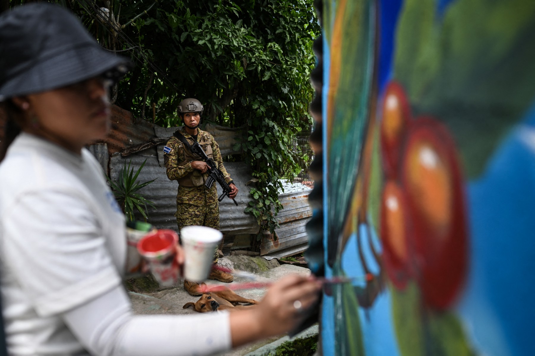 Um soldado salvadorenho faz a guarda na comunidade 10 de Octubre, em San Marcos, El Salvador, enquanto a artista Elizabeth Guardado pinta o mural "Identidade", em 2 de outubro de 2026.