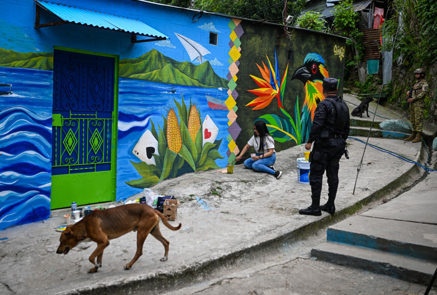 Um soldado salvadorenho faz a guarda na comunidade 10 de Octubre, em San Marcos, El Salvador, enquanto a artista Elizabeth Guardado pinta o mural "Identidade", em 2 de outubro de 2026.