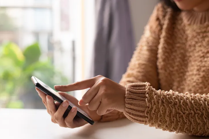 Close-up of girl in sweater viewing online information on smartphone. Unrecognizable lady sitting at table and using modern gadget. Technology concept